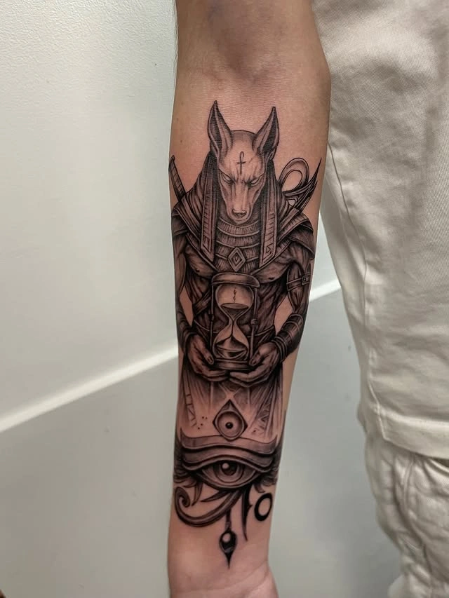 Tatuaj pe braț cu figura lui Anubis ținând o clepsidră, detalii geometrice și ochi stilizat.