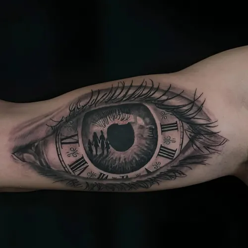Tatuaj alb-negru pe antebraț reprezentând un ochi realist, cu iris detaliat, gene fine, cifre romane de ceas în jurul pupilei și siluete reflectate în interior.