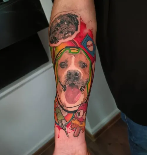 Tatuaj mare pe antebraț, cu două portrete de câini integrate într-o compoziție colorată, cu elemente grafice și fundal roșu, verde, galben și albastru.