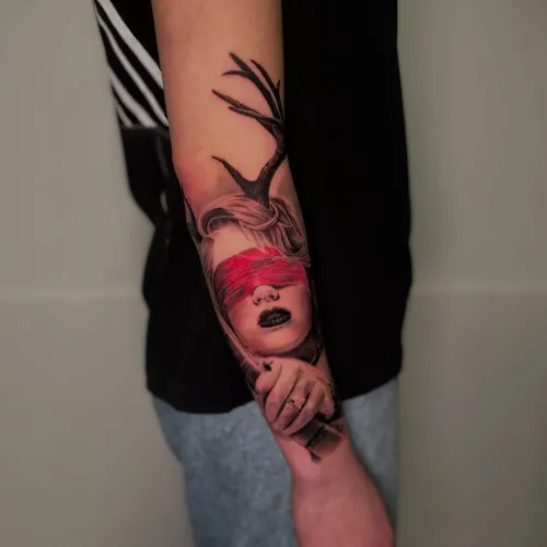 Tatuaj pe antebraț cu portret feminin realist, legat la ochi cu o bandă roșie, elemente negre ca ramuri deasupra capului și o mână detaliată în partea de jos.