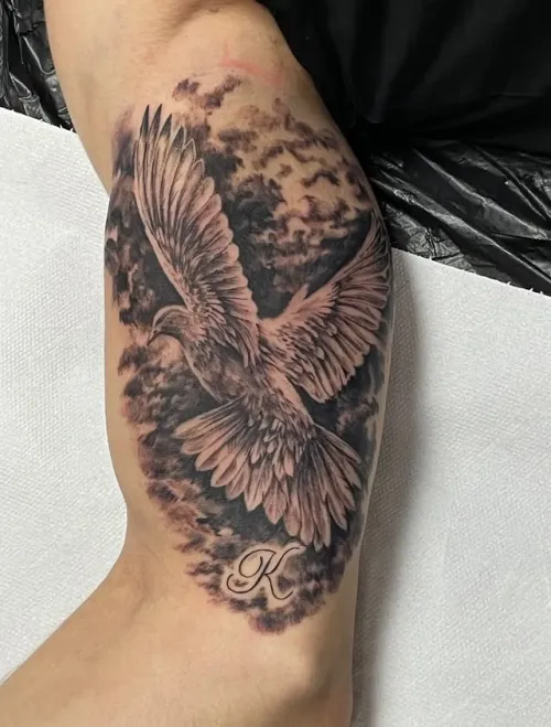 Tatuaj mare pe gambă reprezentând o pasăre cu aripile deschise, realizată în negru și gri, cu fundal fumuriu și inițiala K dedesubt.