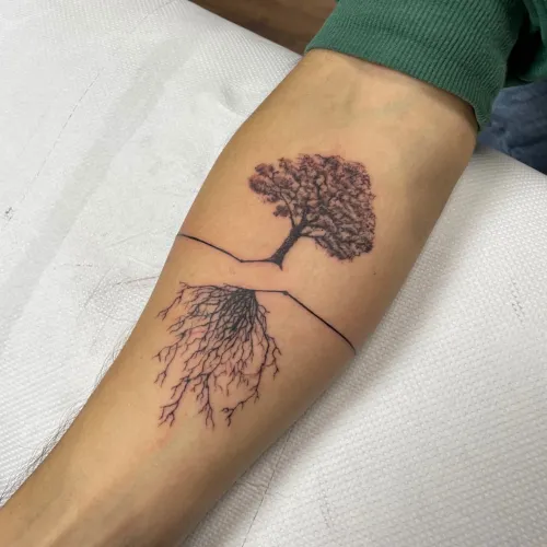 Tatuaj negru pe antebraț reprezentând un copac detaliat deasupra unei linii fine și rădăcini ramificate sub linie.