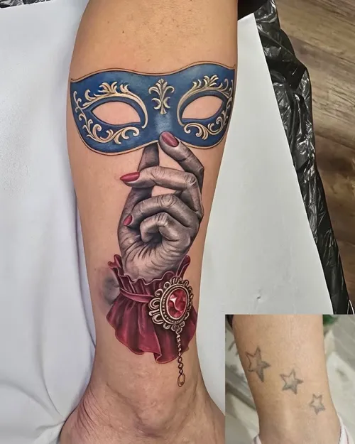 Tatuaj color pe gambă reprezentând o mână realistă cu unghii roșii care ține o mască venețiană albastră cu ornamente aurii, completată de o manșetă roșie și bijuterie decorativă.